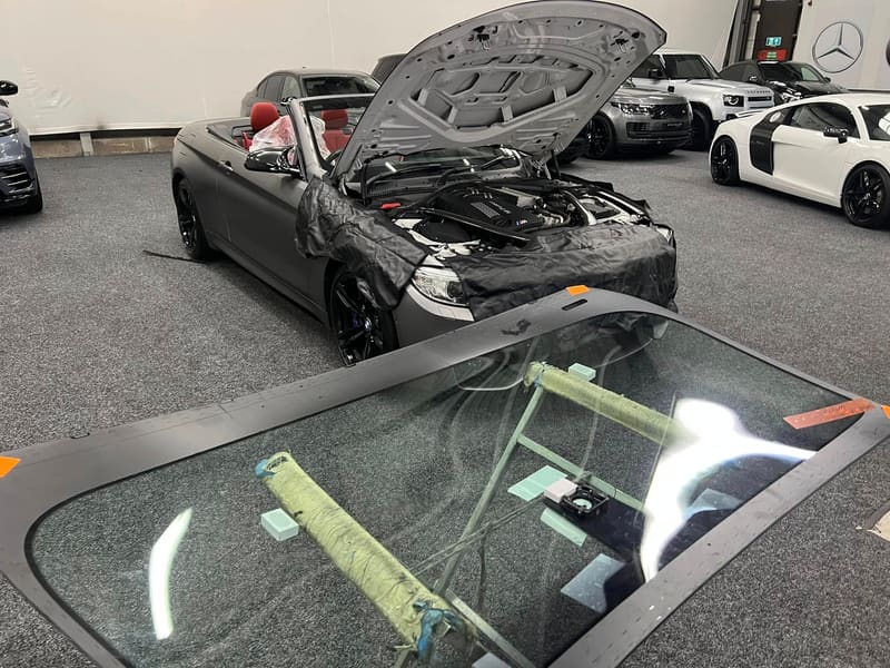 A windscreen laid out on a stand beside a BMW M4 convertible with its screen removed and the bodywork covered